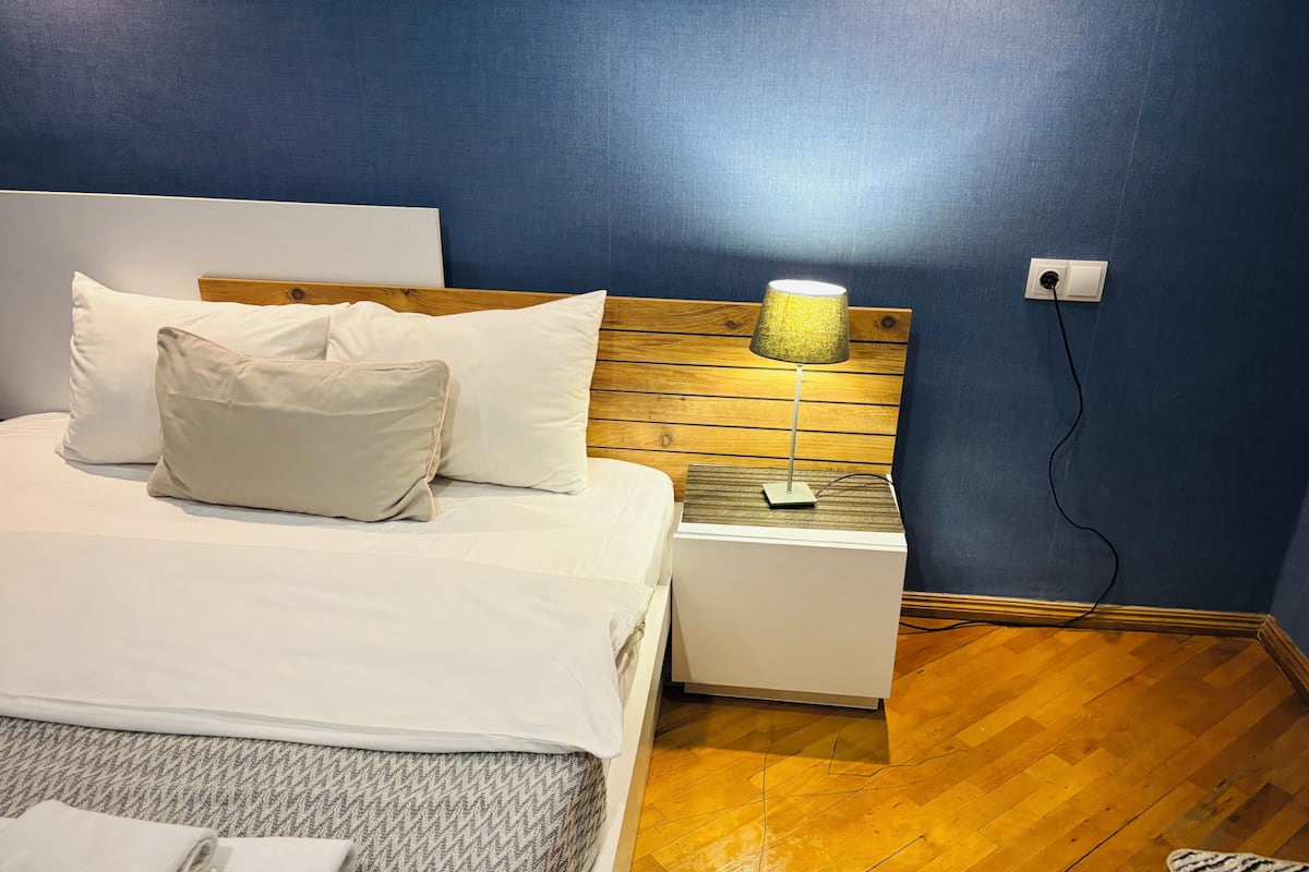A neatly made bed with pillows, a bedside lamp, and a power outlet on the wall.