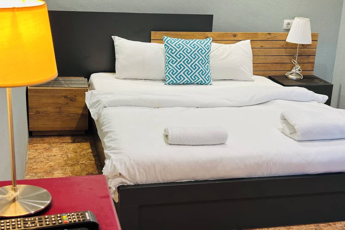 A hotel room with a bed, two pillows, a nightstand with a lamp, a remote control, and a red table.