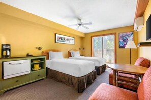 Quarto de hotel com duas camas, um aparador verde, um frigorífico pequeno, um ventilador de teto e uma janela com vista.