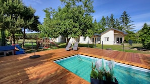 Une terrasse en bois avec une piscine, une aire de jeux et une maison en arrière-plan.
