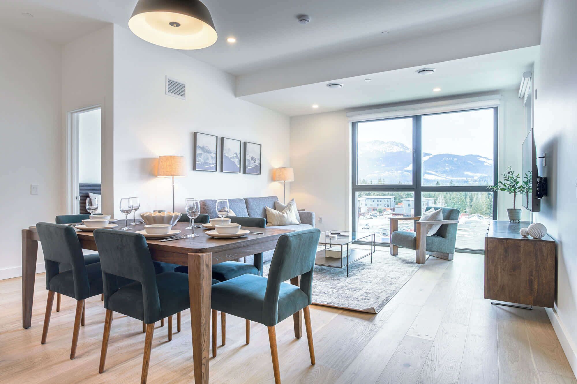 A modern dining area with a wooden table, dark chairs, and a large window offering a mountain view.