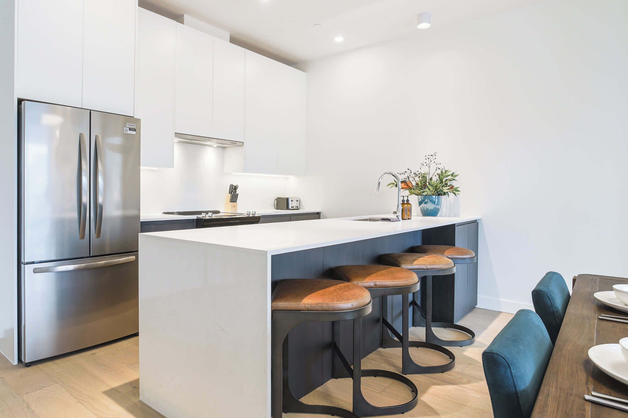 A modern kitchen with a stainless steel refrigerator, a central island with stools, and a dining area with a table and chairs.