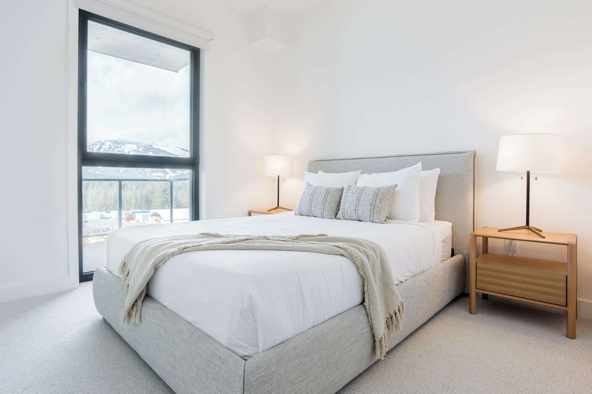 A bedroom with a large window, a bed with white linens, two bedside tables with lamps, and a view of a snowy landscape.