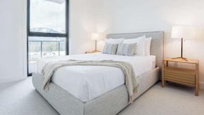 Une chambre avec une grande fenêtre, un lit recouvert de draps blancs, deux tables de chevet avec des lampes, et une vue sur un paysage enneigé.