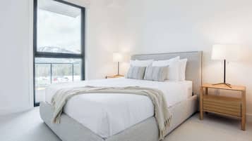 A bedroom with a large window, a bed with white linens, two bedside tables with lamps, and a view of a snowy landscape.