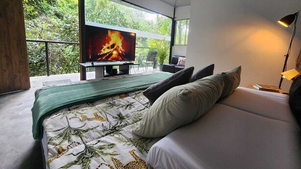 Un dormitorio con una televisión grande, una cama con colcha verde y blanca, una lámpara de noche y vistas al exterior con vegetación.