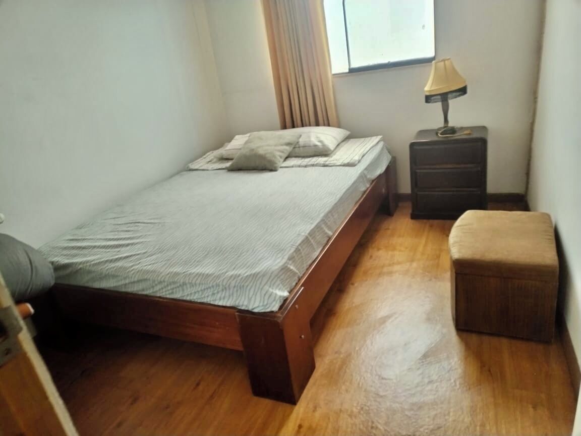 Une petite chambre avec un lit simple, un plancher en bois, une table de chevet avec une lampe et une fenêtre avec des rideaux.