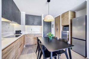Eine moderne Küche mit dunklen Schränken, einer Marmorarbeitsplatte, einem Esstisch mit Stühlen, einer Pendelleuchte und einem Edelstahl-Kühlschrank.