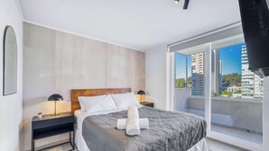 A modern hotel room with a large bed, a nightstand, a lamp, and a view of the city through a large window. - Modern Coast Concón- 2D w/ Wifi (Concón)