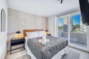 Ein modernes Hotelzimmer mit einem großen Bett, einem Nachttisch, einer Lampe und Blick auf die Stadt durch ein großes Fenster.