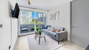 A modern living room with a grey sofa, a coffee table with plants, a flat-screen TV mounted on the wall, and a large window offering a city view. - Modern Coast Concón- 2D w/ Wifi (Concón)