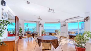 A spacious room with a wooden floor, round tables with brown tablecloths, wicker chairs, potted plants, and large windows offering a view of the sea. - Vista Pacífico- 2D , Sea View and Wifi (Viña del Mar)