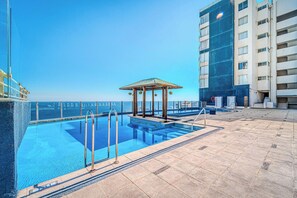 A rooftop pool with a gazebo, clear sky, and a high-rise building in the background.