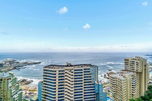 A high-rise hotel with multiple balconies overlooking a coastal area with rocky terrain and the ocean.
