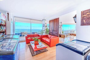 A spacious living area with a red sofa, a glass coffee table, and a view of the sea through large windows.