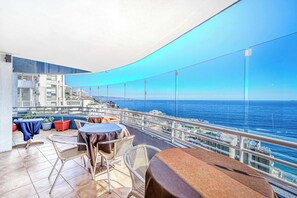Ein Balkon mit Tischen und Stühlen, mit Blick auf eine Küstenstadt und das Meer.