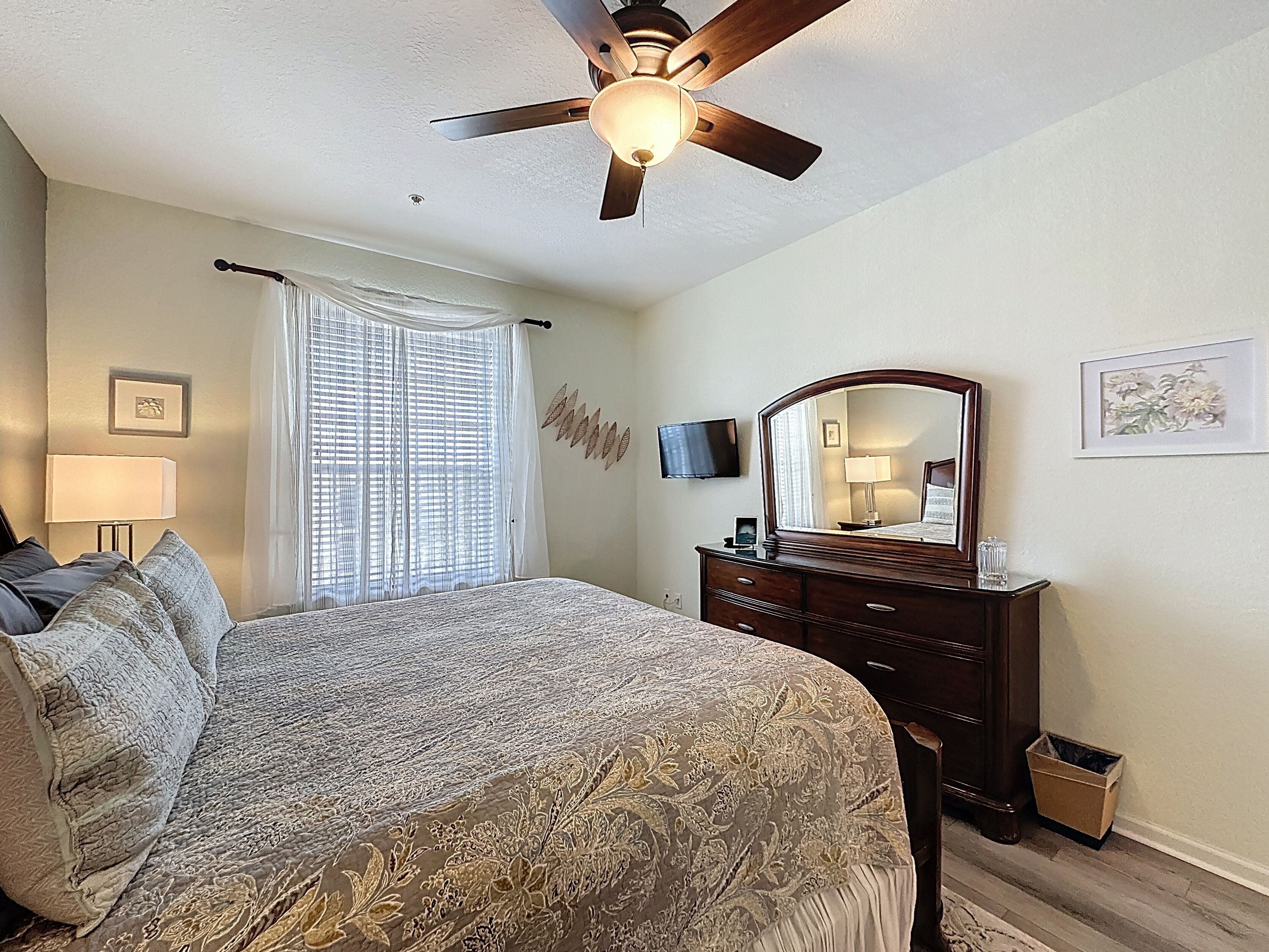 A bedroom with a bed, a ceiling fan, a dresser, a mirror, a TV, and a window with curtains.