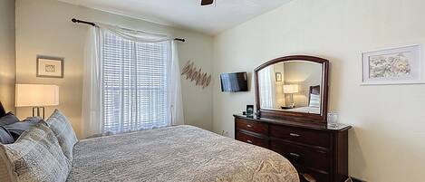 A bedroom with a bed, a ceiling fan, a dresser, a mirror, a TV, and a window with curtains.