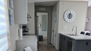 A modern kitchen with a clock, grey sofas with patterned cushions, and a wooden floor. - Welcome to vacation Home five minutes from the beach. 🏖️ (Pensacola)