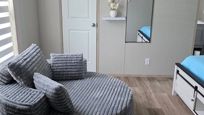 A room with a round, cushioned seating area, a bed with a blue mattress, a white door, a mirror, and a small shelf with flowers. - Welcome to vacation Home five minutes from the beach. 🏖️ (Pensacola)