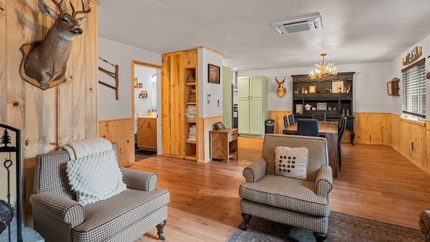 A cozy living room with wooden flooring, two patterned armchairs, a mounted deer head, and a dining area with a chandelier. - The Hideaway (Big Bear Lake)