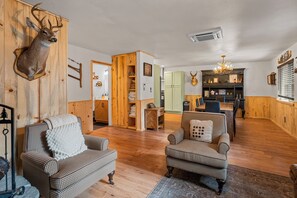 A cozy living room with wooden flooring, two patterned armchairs, a mounted deer head, and a dining area with a chandelier. - The Hideaway (Big Bear Lake)