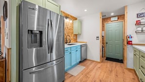 A compact kitchen with a stainless steel refrigerator, light blue cabinets, a wooden backsplash, and a green door marked 'EXIT'. - The Hideaway (Big Bear Lake)