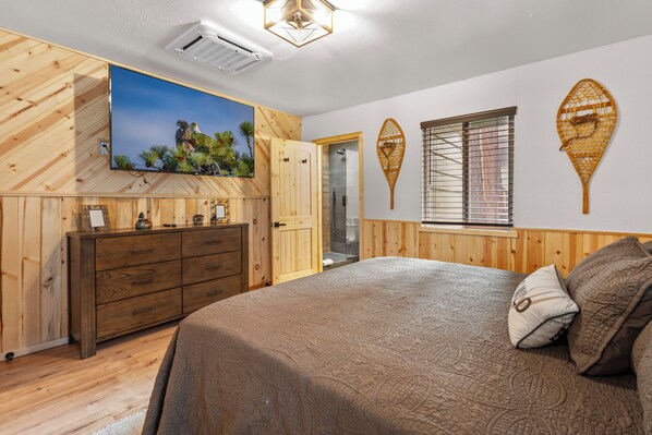 A bedroom with a large bed, wooden paneling, a television, and a shower. - The Hideaway (Big Bear Lake)