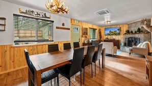 A wooden dining area with a large table and chairs, a chandelier, and a fireplace in the background. - The Hideaway (Big Bear Lake)