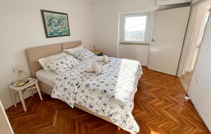 Ein Schlafzimmer mit Bett, Nachttisch, einem Gemälde, einem Fenster und einer Tür.