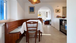Zona pranzo con una panca, sedie in legno, un tavolo con tovaglia bianca, un lampadario in vimini e angolo cottura con frigorifero.