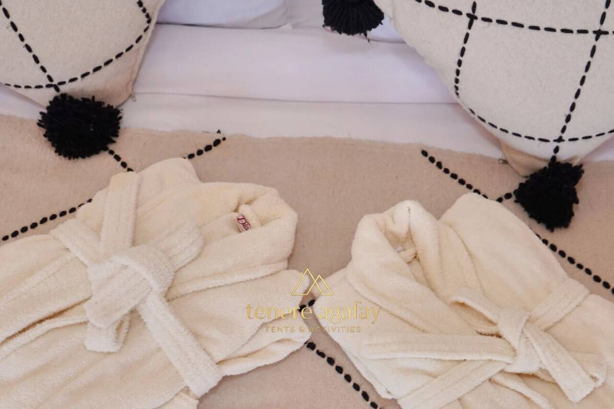 Dos albornoces blancos con borlas negras sobre un suelo color beige, atados en lazo, con un logotipo y el texto 'tenere agafay tents & activities' en el centro.