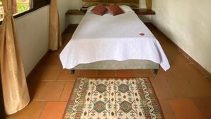 A small, narrow room with a single bed, a window with curtains, a wooden ceiling, and a patterned rug.