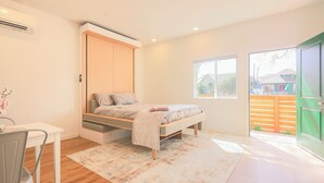 Ein Schlafzimmer mit Bett, Schreibtisch, Stuhl, Fenster und einer Tür zu einem Außenbereich.