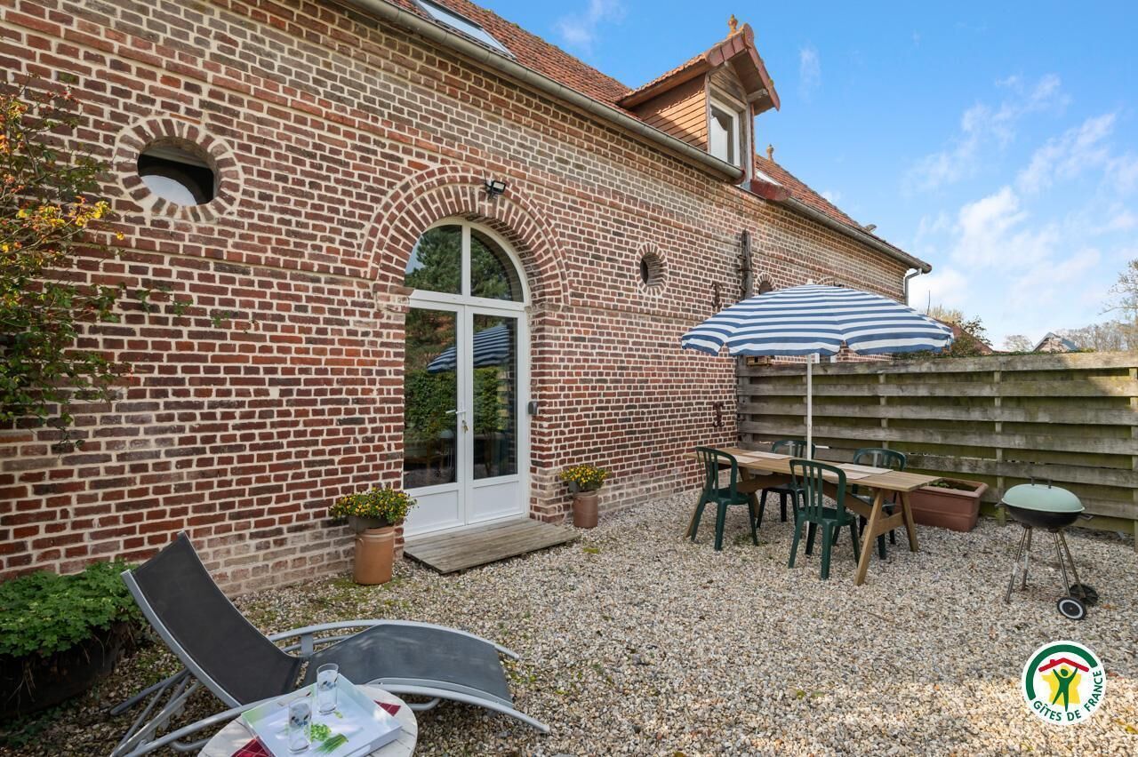A brick building with a patio area featuring a table and chairs, a grill, and potted plants.