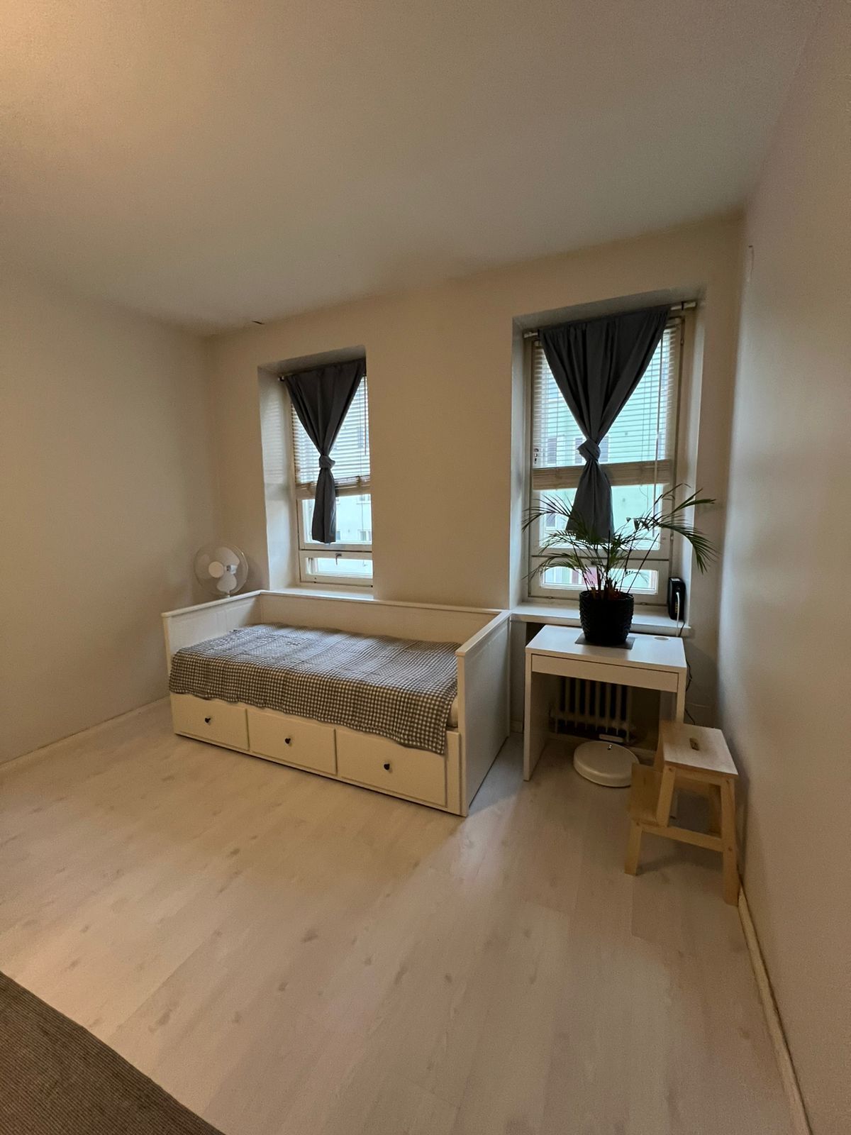 Um pequeno apartamento de um quarto, com cama, escrivaninha, duas janelas com cortinas, radiador e um banquinho.