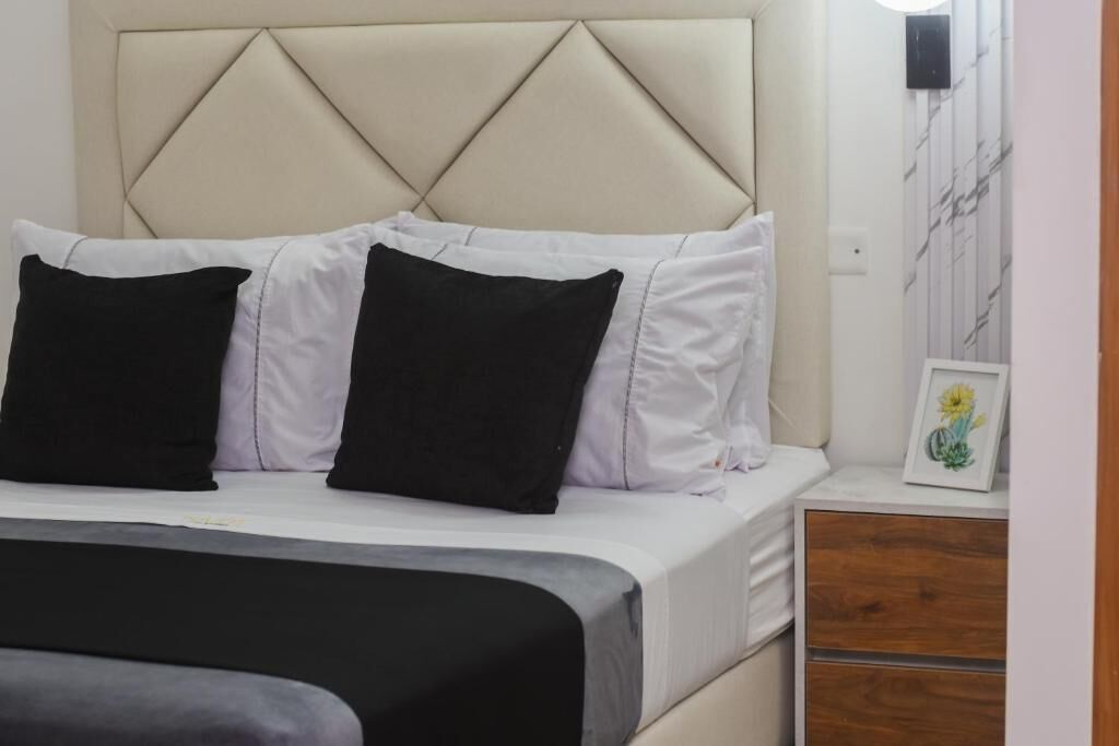 Una cama con cabecera, almohadas blancas y negras, una mesita de noche de madera con un cuadro enmarcado y una mesita auxiliar.