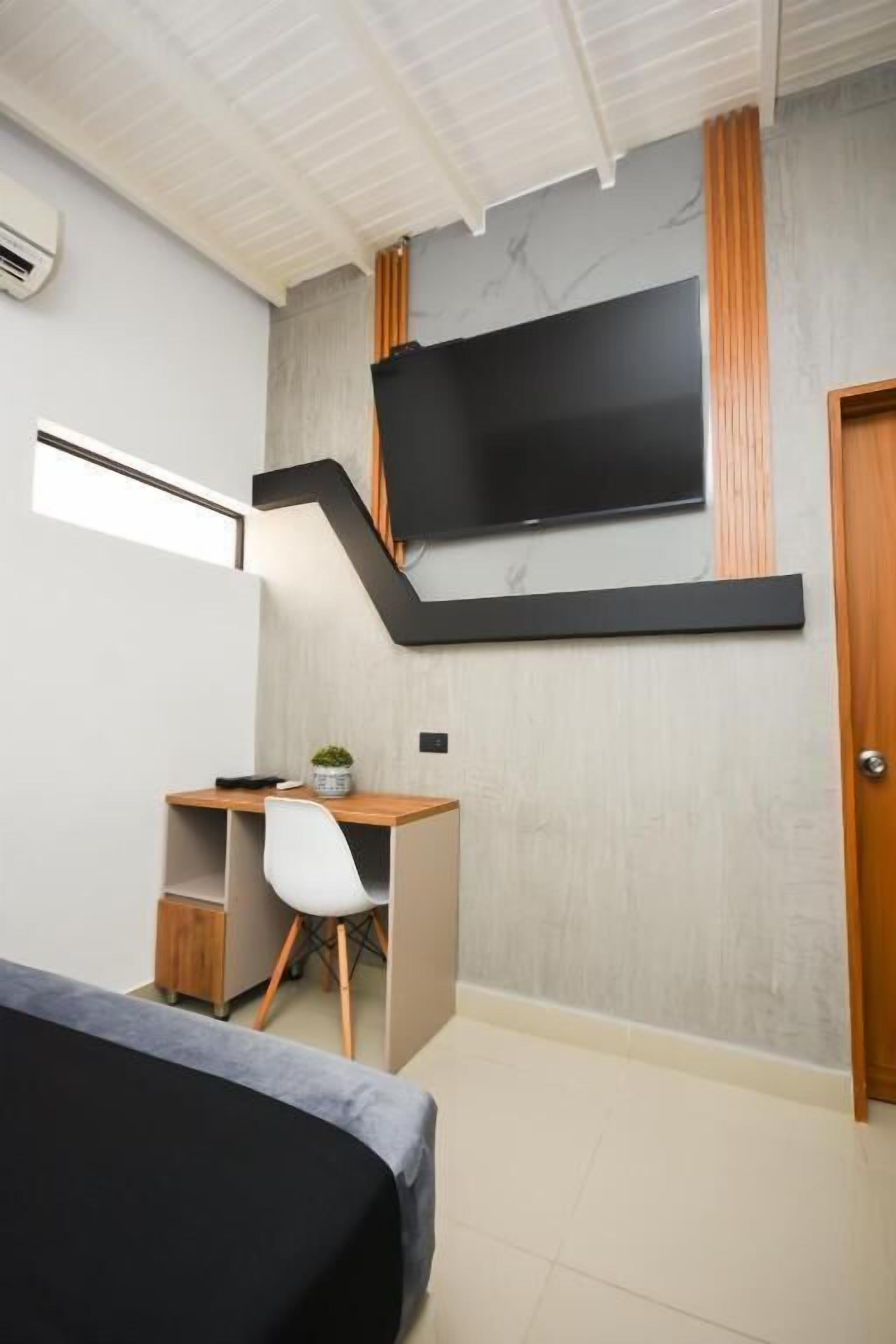 Una habitación moderna con una cama, un escritorio con silla, un televisor montado en la pared y una puerta.