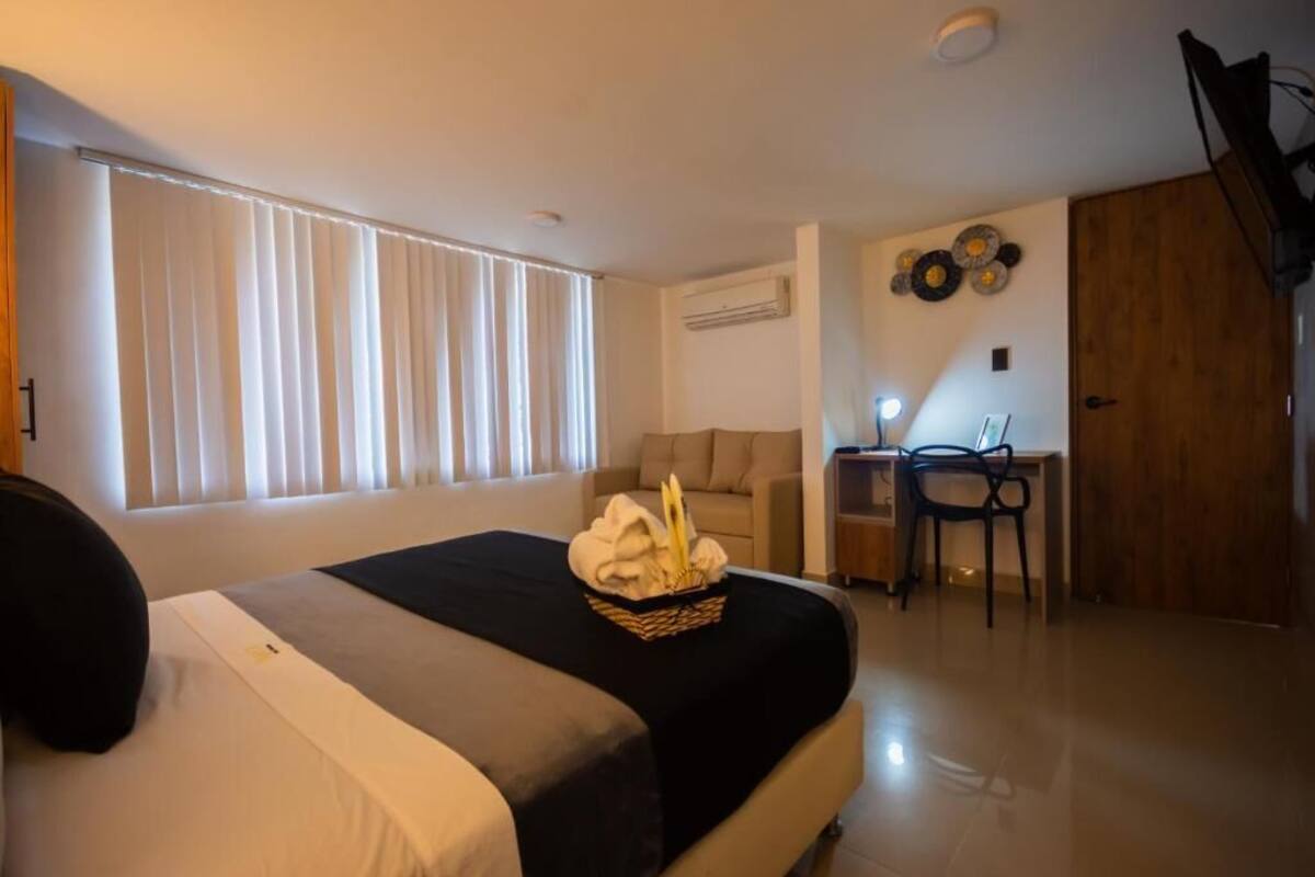 Quarto de hotel com cama, escrivaninha e cadeira. Há uma janela com persianas, ar condicionado fixado na parede e uma porta.