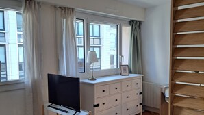 Habitación con una televisión sobre un mueble bajo, un aparador blanco, una mesita y una ventana con cortinas.