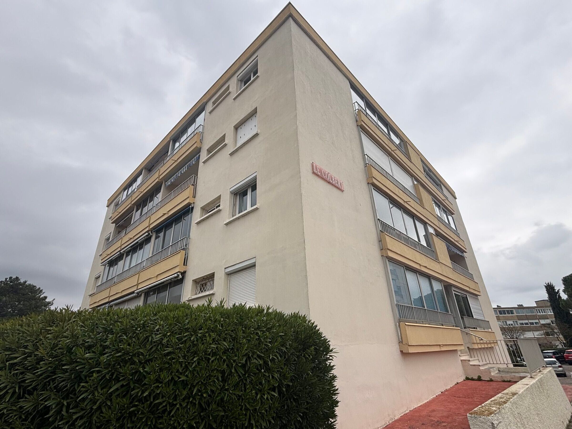 Un edificio a più piani con balconi, un cartello con la scritta 'LE COLBERT' e un siepe in primo piano.