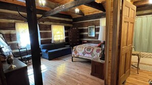 A log cabin interior with a wooden floor, a bed with a patterned bedspread, a dresser, a window with curtains, and a door leading to another room. - Blackberry Inn at Heritage Farm (Huntington)