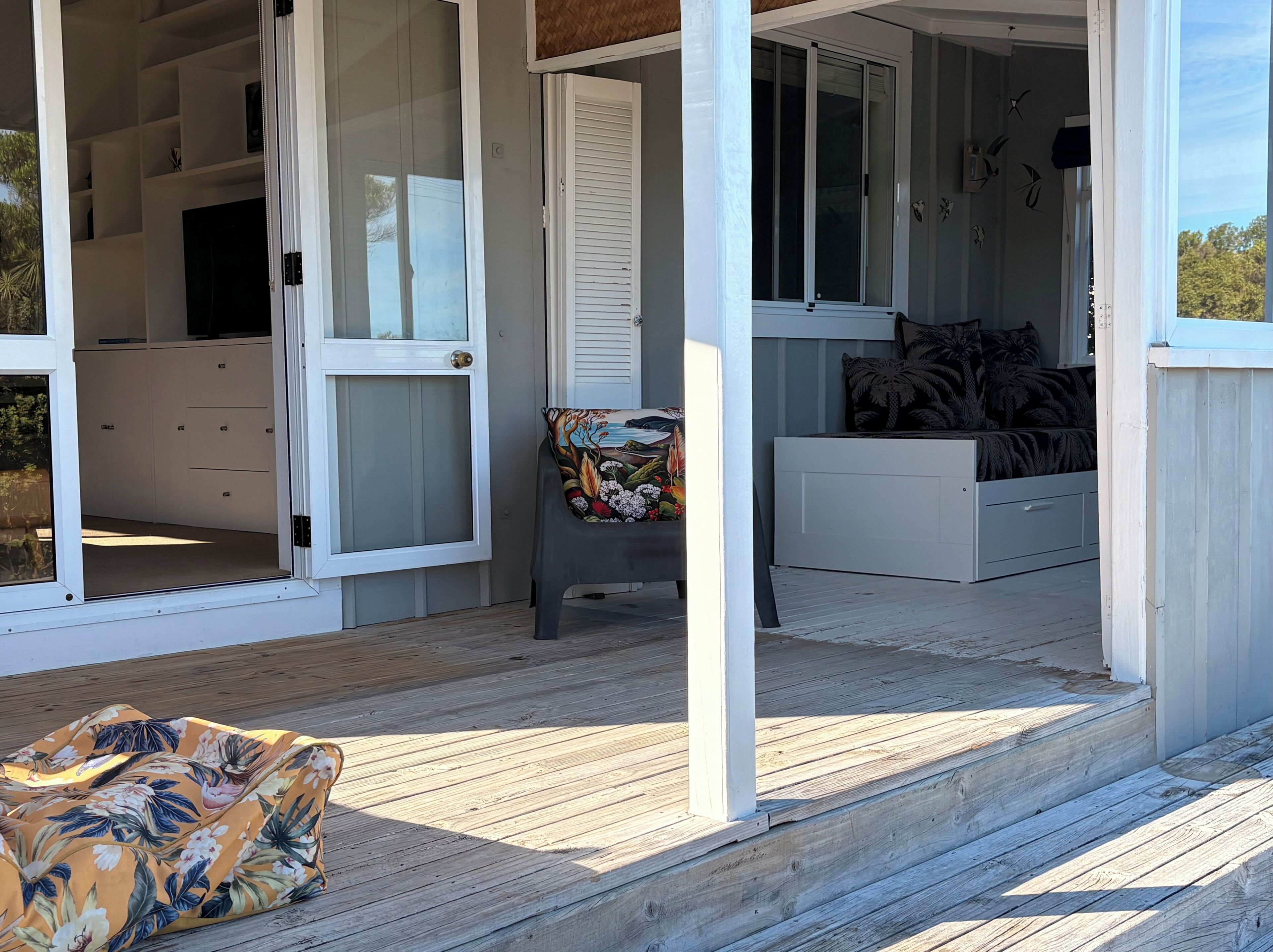 Uma área externa coberta com deck de madeira, um banco com almofada estampada e uma cama com travesseiro padronizado.