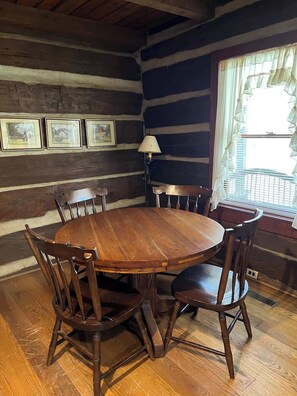 A rustic wooden dining room with a round table and chairs, framed pictures on the wall, and a window with curtains.