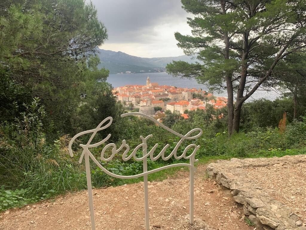 A sign with 'Korsula' written on it, surrounded by greenery and overlooking a coastal town with red-roofed buildings.