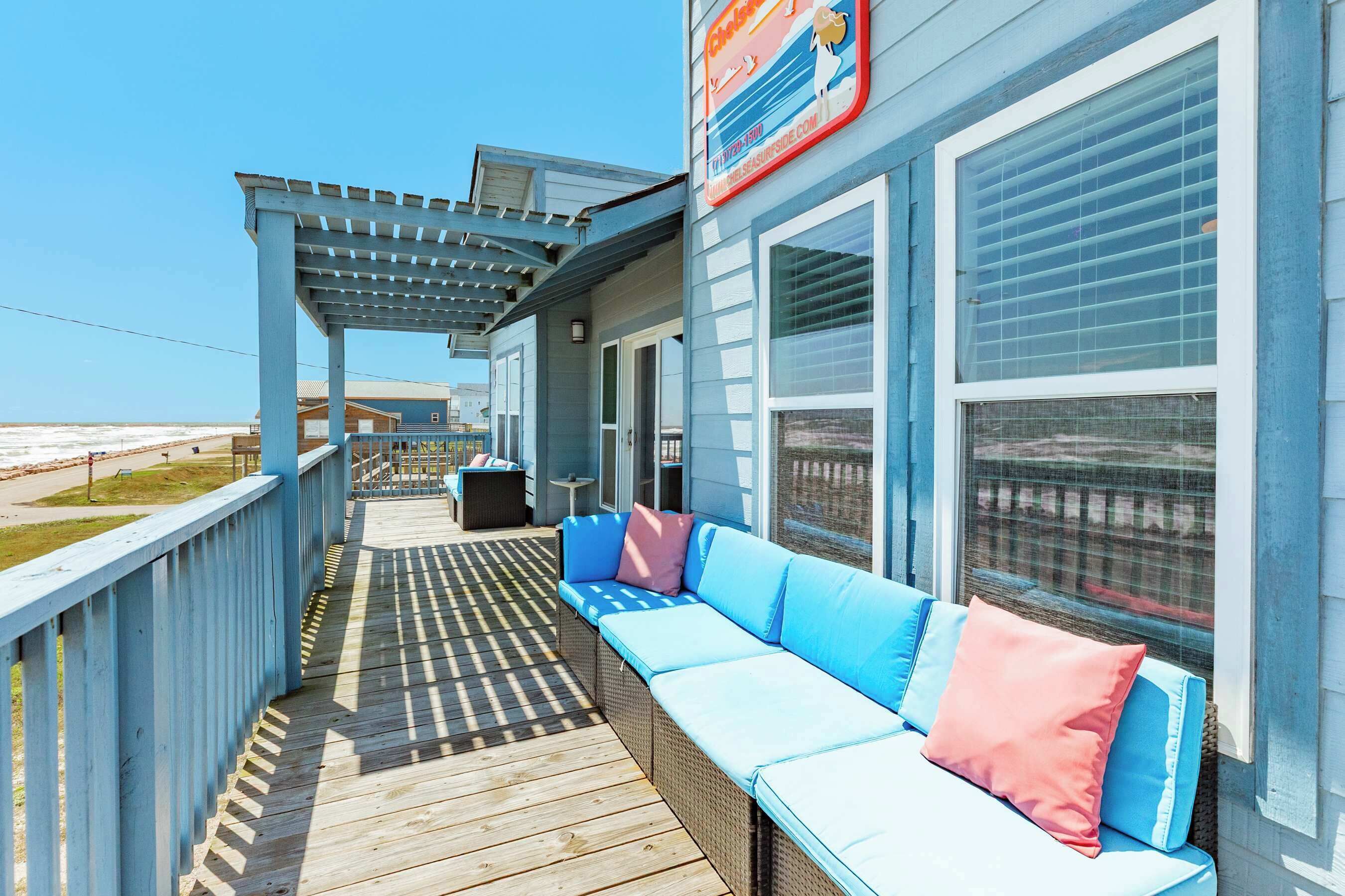 A covered deck with a wicker sofa and a pergola, overlooking a beach.