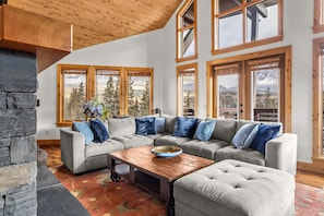 A spacious living room with a stone fireplace, a large sectional sofa, and multiple windows offering mountain views. - Luxury Penthouse Unparalleled Views & Comfort (Canmore)