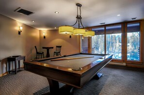 A pool table with a hanging light fixture, two chairs, and a view of trees through large windows. - Luxury Penthouse Unparalleled Views & Comfort (Canmore)