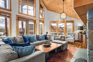 A spacious living room with a large sofa, armchairs, a wooden coffee table, and a chandelier. Large windows offer a view of a mountain landscape. - Luxury Penthouse Unparalleled Views & Comfort (Canmore)