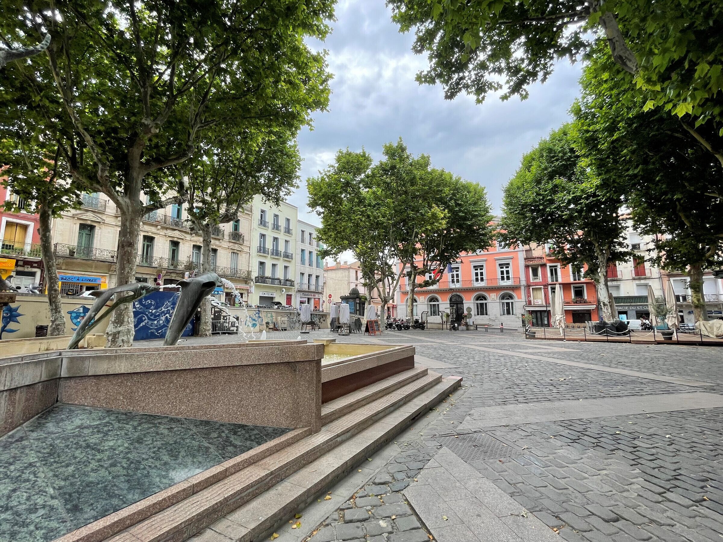 Une place publique avec une fontaine, pavée de pierres, entourée d’arbres et de bâtiments dotés de balcons.